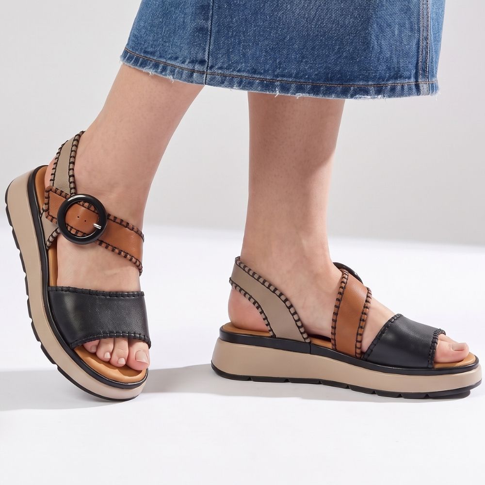 A lady in a denim skirt wearing a pair of sandals with black, taupe and tan leather straps with stitched edged detailing and a round buckle, set on a beige platform sole, standing against a studio background.