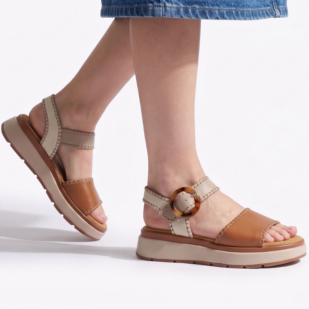 A lady in a denim skirt wearing a pair of sandals with tan, taupe and cream leather straps with stitched edged detailing and a round buckle, set on a beige platform sole, standing against a studio background.
