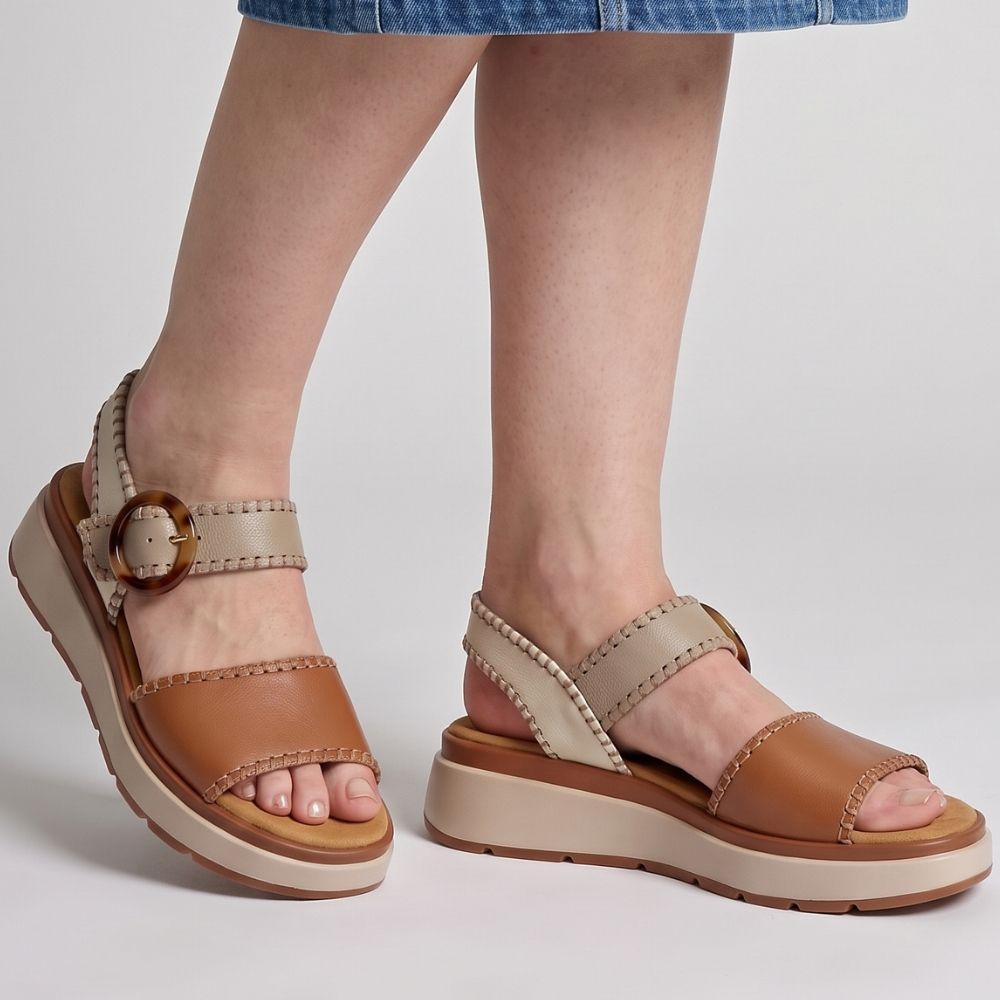 A lady in a denim skirt wearing a pair of sandals with tan, taupe and cream leather straps with stitched edged detailing and a round buckle, set on a beige platform sole, standing against a studio background.