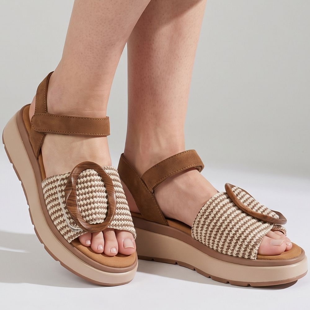 A lady wearing a pair of sandals with a tan and cream woven strap across the vamp showcasing a statement oversized buckle, tan ankle straps and a beige platform sole against a studio background.