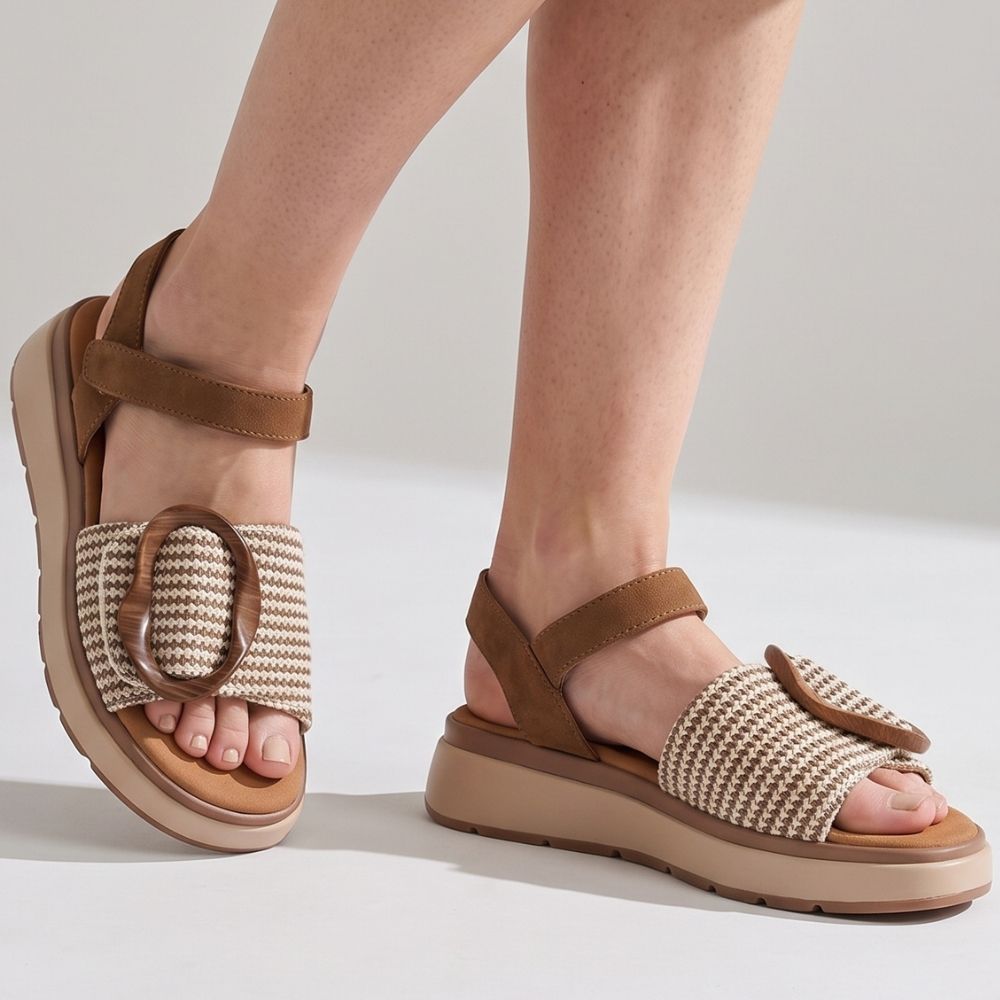 A lady wearing a pair of sandals with a tan and cream woven strap across the vamp showcasing a statement oversized buckle, tan ankle straps and a beige platform sole against a studio background.