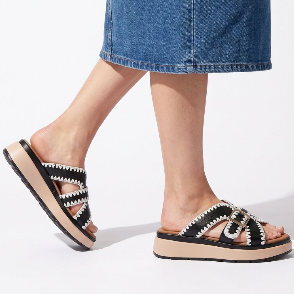 A lady wearing a denim skirt and a pair of black leather slides with white stitched edging, a bronze buckle and a brown and black platform sole, set against a white background.