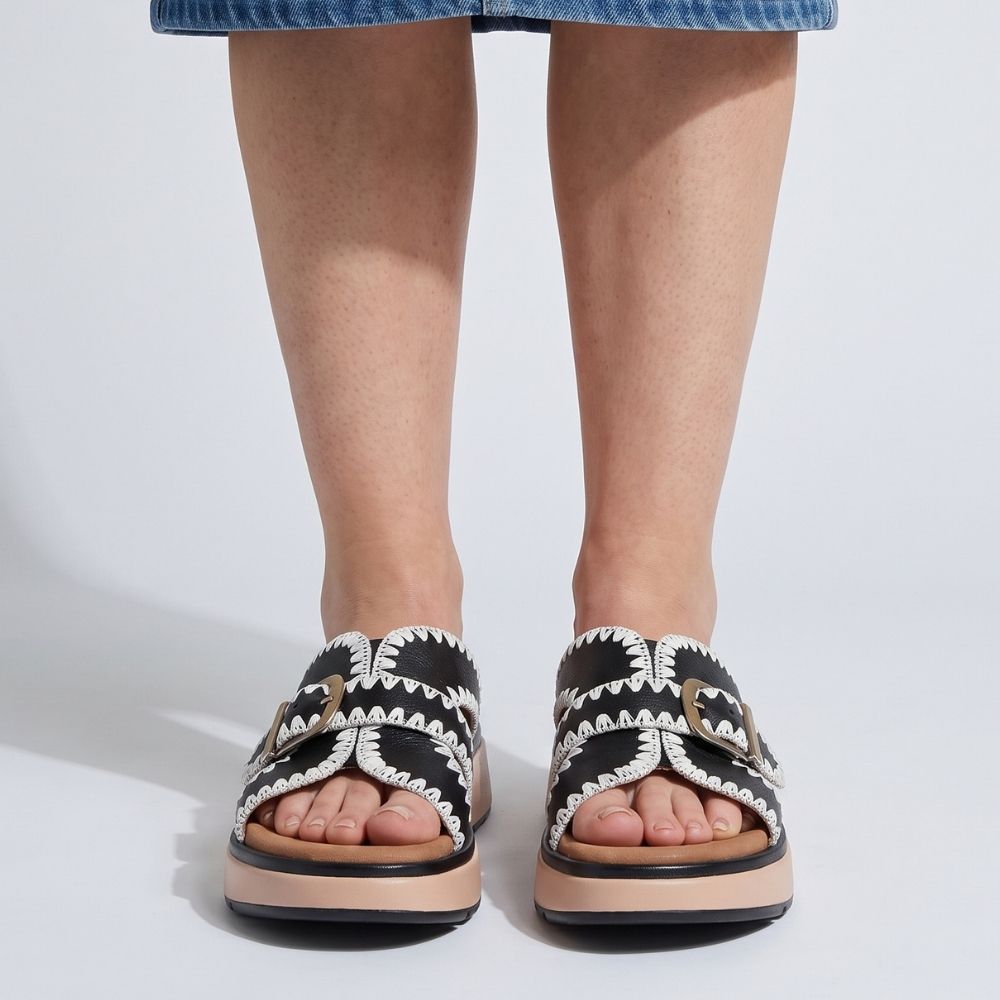 A lady wearing a denim skirt and a pair of black leather slides with white stitched edging, a bronze buckle and a brown and black platform sole, set against a white background.