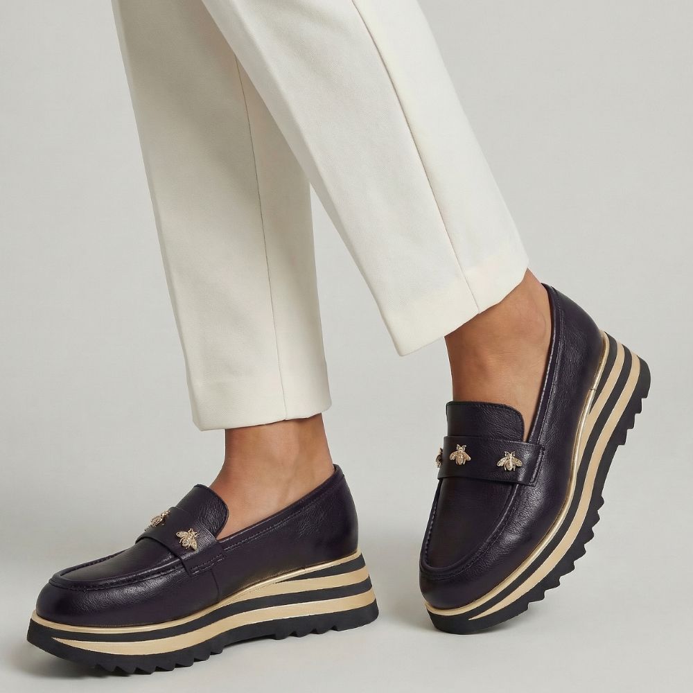 Black loafers with gold bees and gold stripes worn with white pants on a light gray background