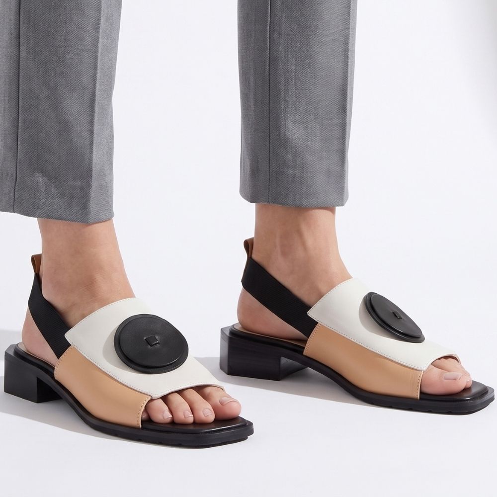 A pair of women's block-heel sandals worn with gray trousers. The sandals feature a color-blocked design with black straps and soles, a beige toe strap, and a cream upper panel decorated with a large black circular disc.