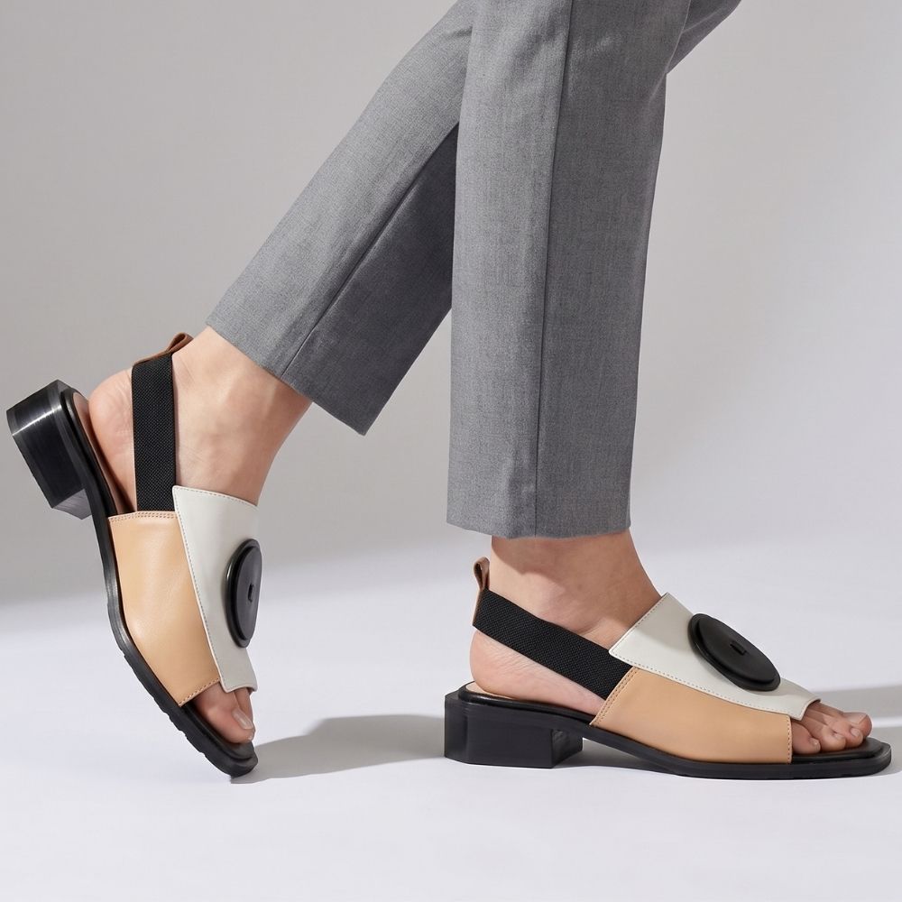 A person wearing slide sandals with a low block heel, featuring a color-blocked design in tan, cream, and black, with a black leather disk detail on the toe straps.