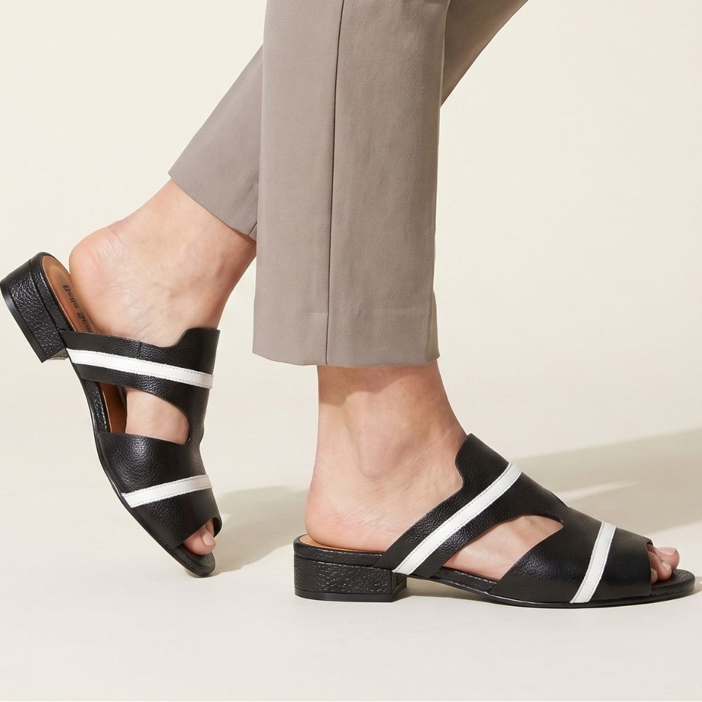 Black leather slide sandals with white stripe accents, shown on a model wearing taupe trousers against a light beige background.