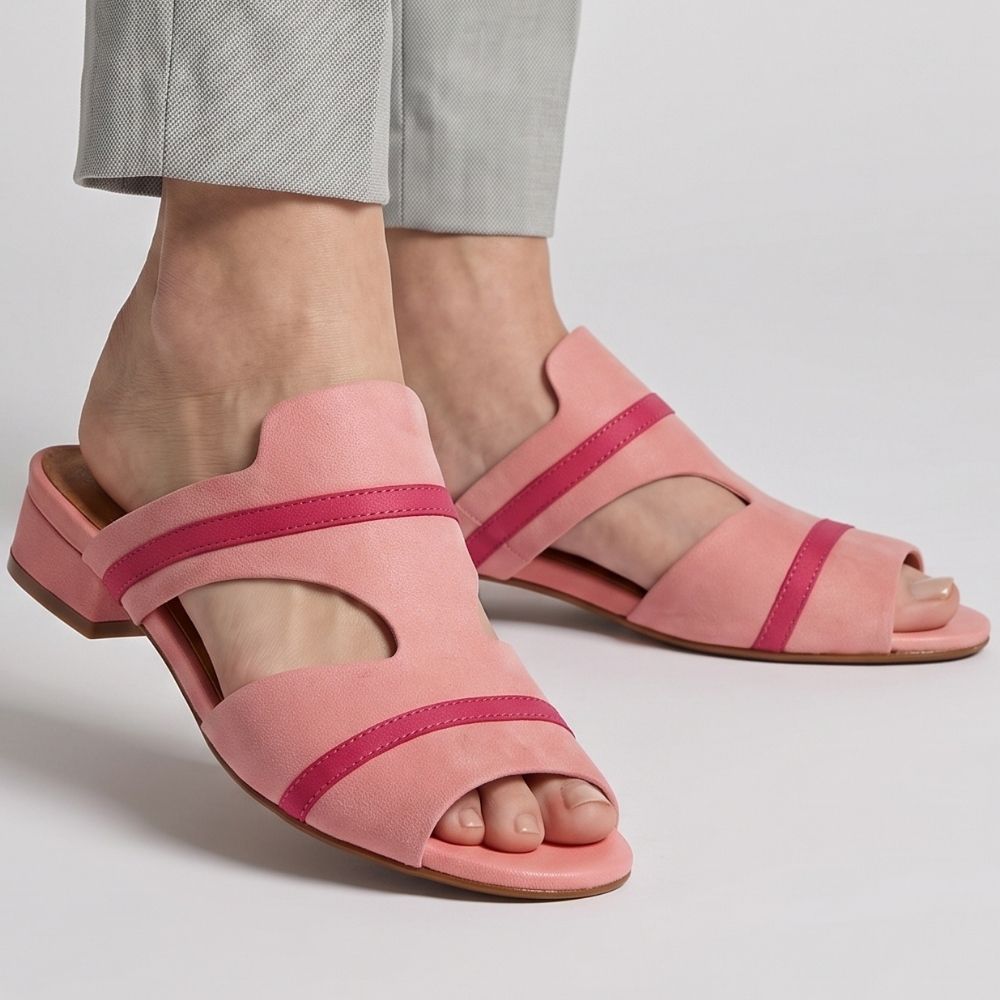 A pair of women's peep-toe slide sandals in pink leather with contrasting magenta-pink straps, shown on a model's feet wearing light gray trousers against a light gray studio background.