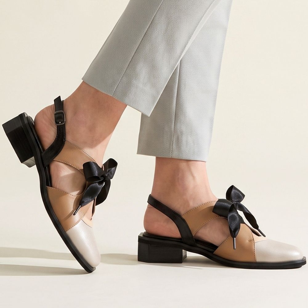 A pair of women's low-heel sandals shown on a model's feet, styled with light gray trousers. The sandals feature a beige and taupe color-blocked toe, black slingback straps, and a black ribbon bow on the front.