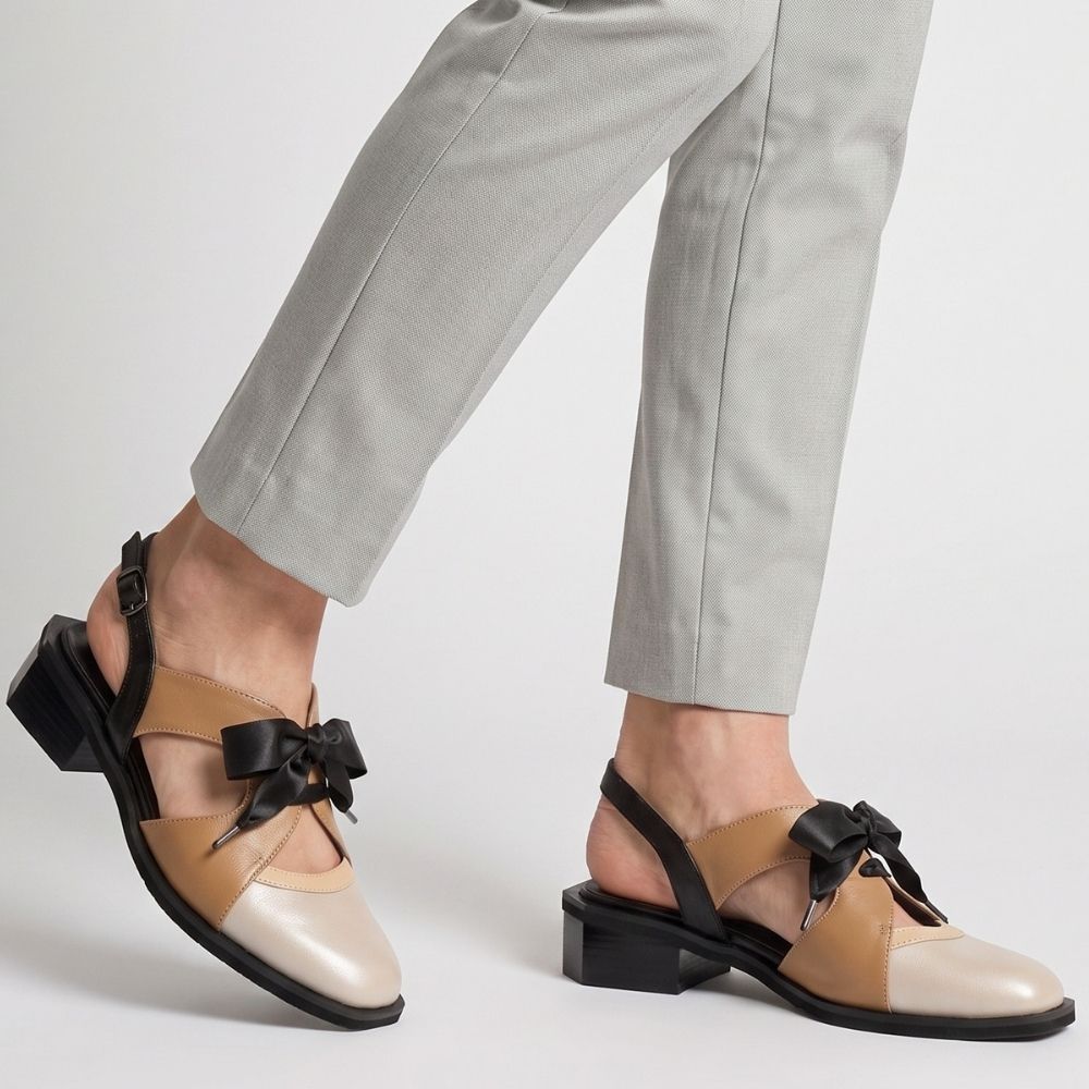 A person wearing light gray cropped trousers and tan-and-taupe low-heel sandals with black bows and black slingback straps, shown against a clean light gray studio background.