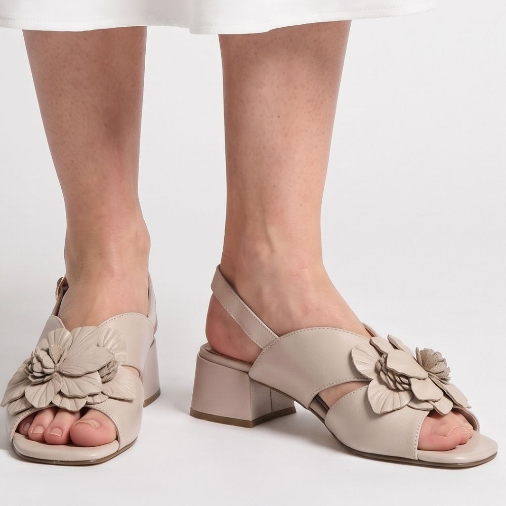 A person wearing a white skirt with a pair of nude leather sandals with a floral leather embellishment across the vamp, peep toe and a low block heel, against a studio background.