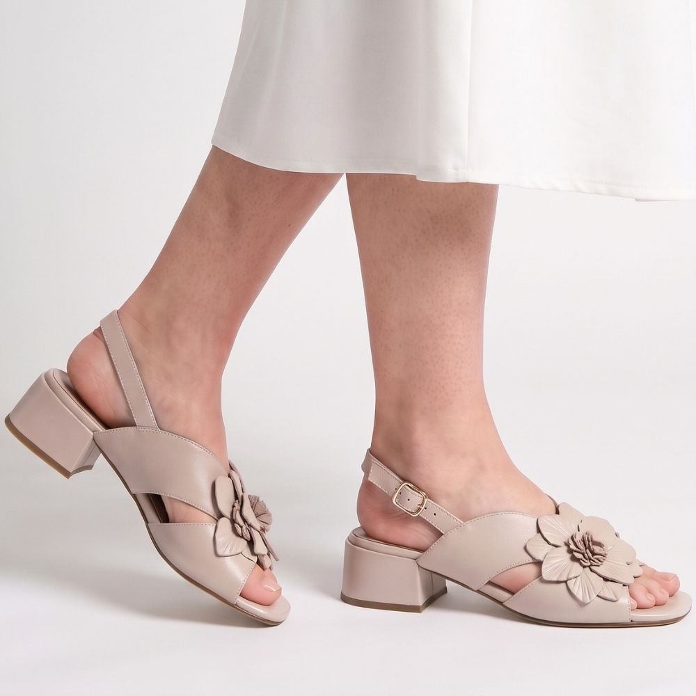 A person wearing a white skirt with a pair of nude leather sandals with a floral leather embellishment across the vamp, peep toe and a low block heel, against a studio background.