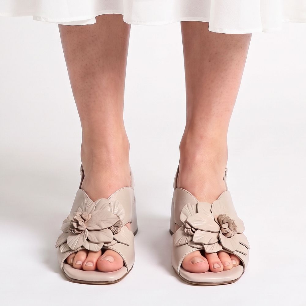 A person wearing a white skirt with a pair of nude leather sandals with a floral leather embellishment across the vamp, peep toe and a low block heel, against a studio background.