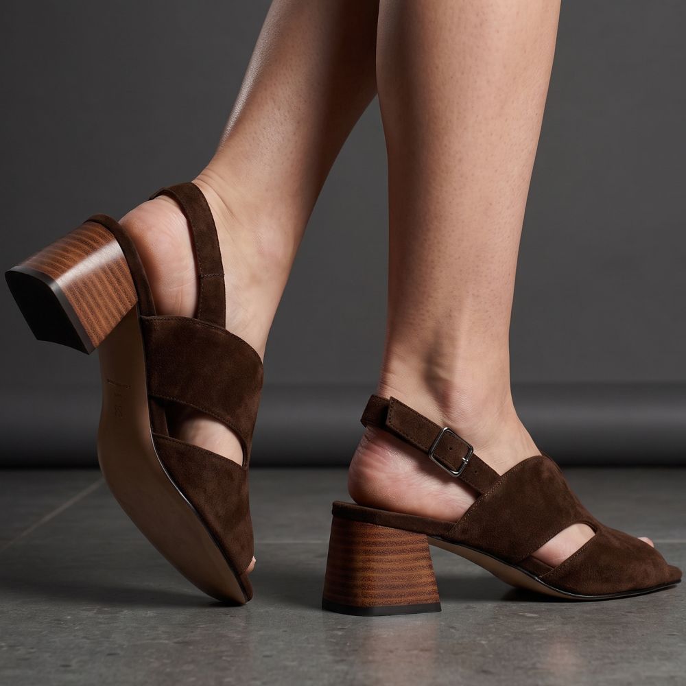 A pair of women's brown suede block-heel sandals with peep toe and cutout side panels, shown on a model's feet against a dark gray studio background.