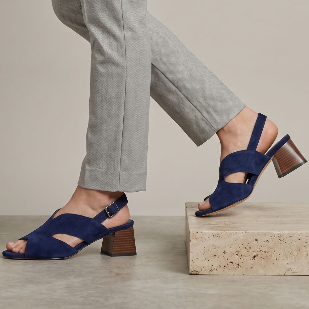 A person wearing navy suede slingback block-heel sandals, paired with light grey trousers, standing on a stone pedestal against a neutral beige background.