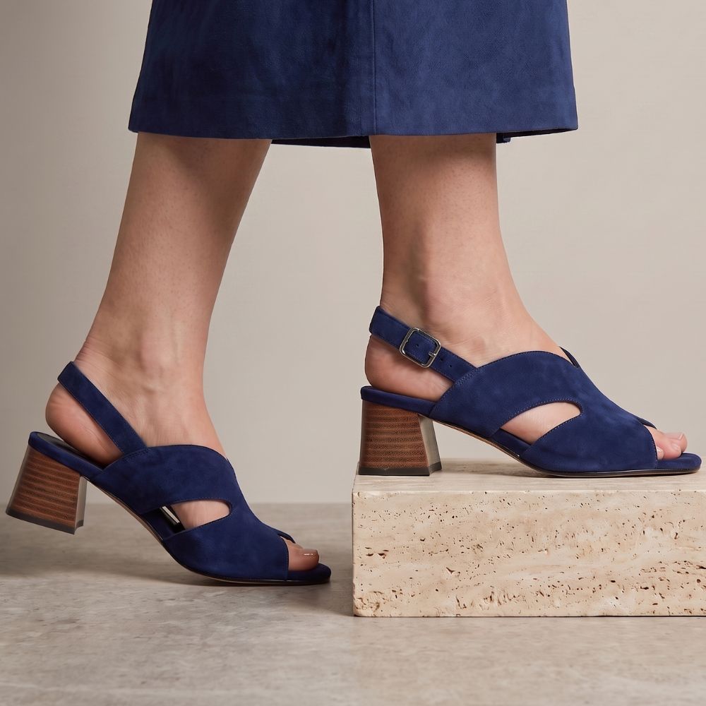 A pair of navy blue suede slingback block-heel sandals with cutout details on the toe and sides, shown on a model's feet. One sandal is placed on a light beige stone block, and the other is on the floor, against a neutral beige background.