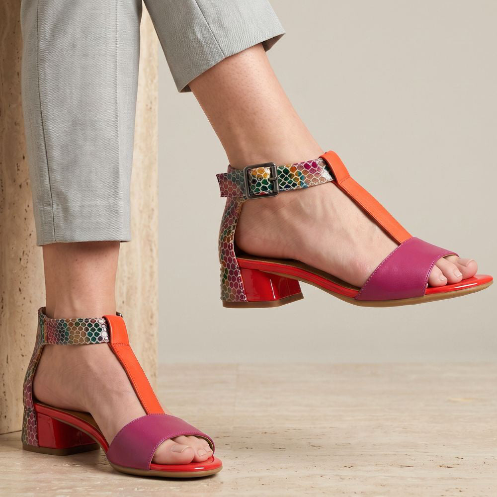 Person with linen pants sitting on a plinth wearing bright pink, orange and multi-coloured T-Strap heels with adjustable buckles and a low heel 