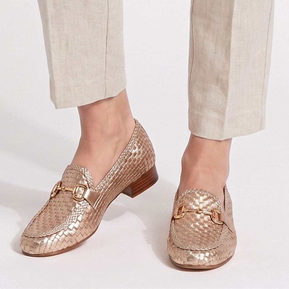 A pair of metallic rose gold woven loafers with gold-tone horsebit details, shown on a model wearing linen trousers against a white studio background.