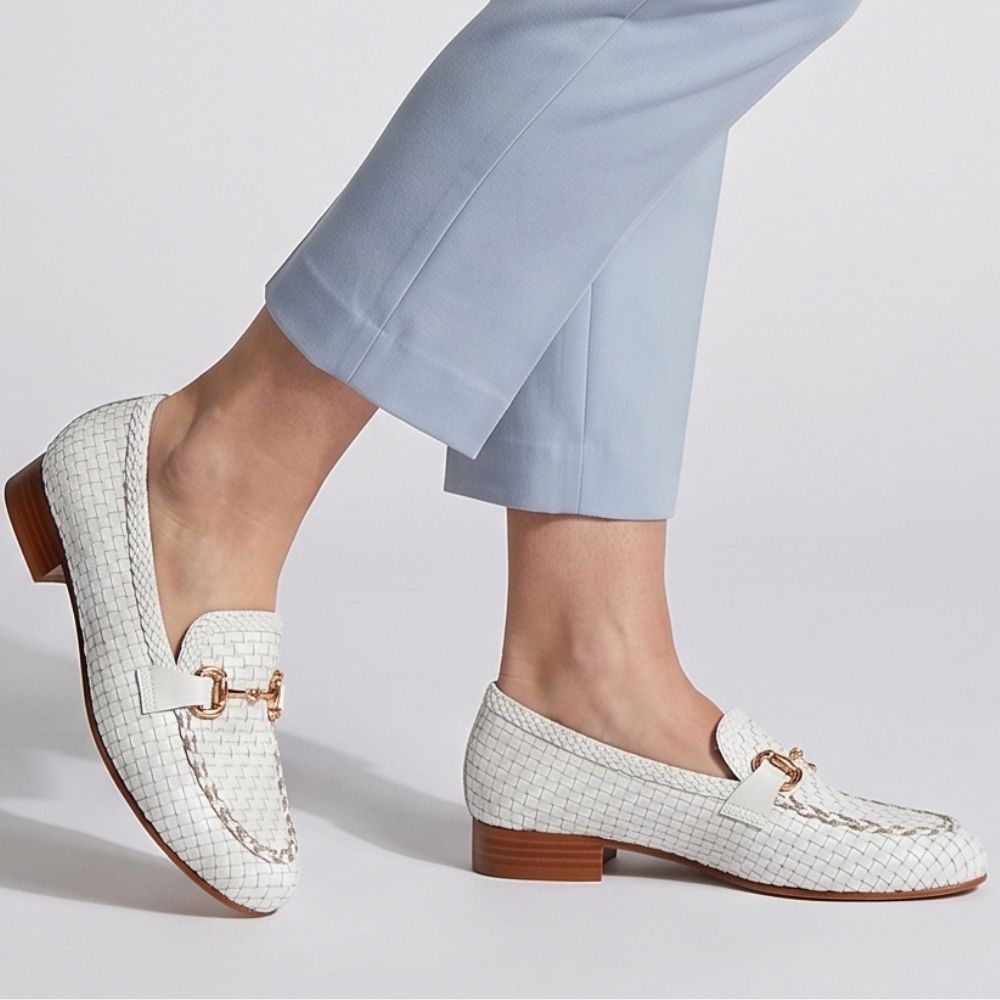 A person wearing white woven leather loafers with gold-tone horsebit details and brown low heels, paired with light blue cropped trousers against a light gray studio background.