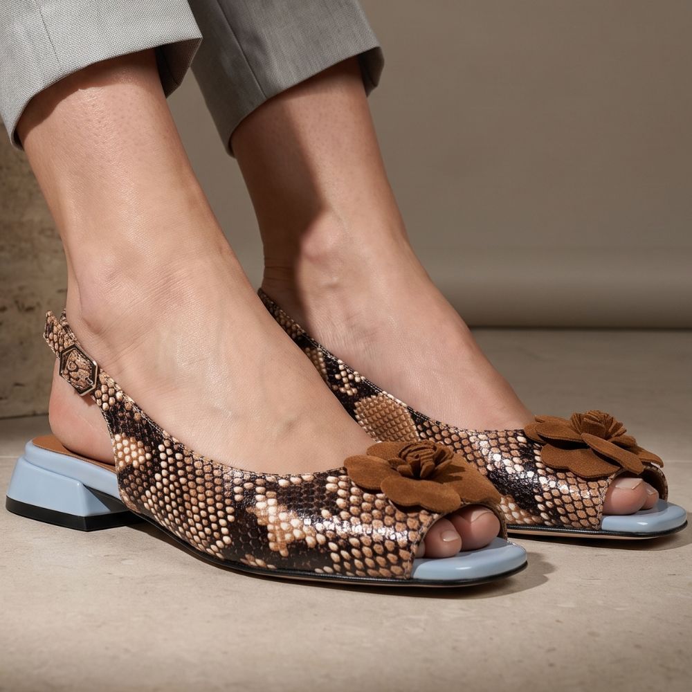 A close-up of a person wearing snakeskin-patterned slingback flat sandals with brown suede flower embellishments on the toes and a light blue block heel, shown against a neutral beige background.