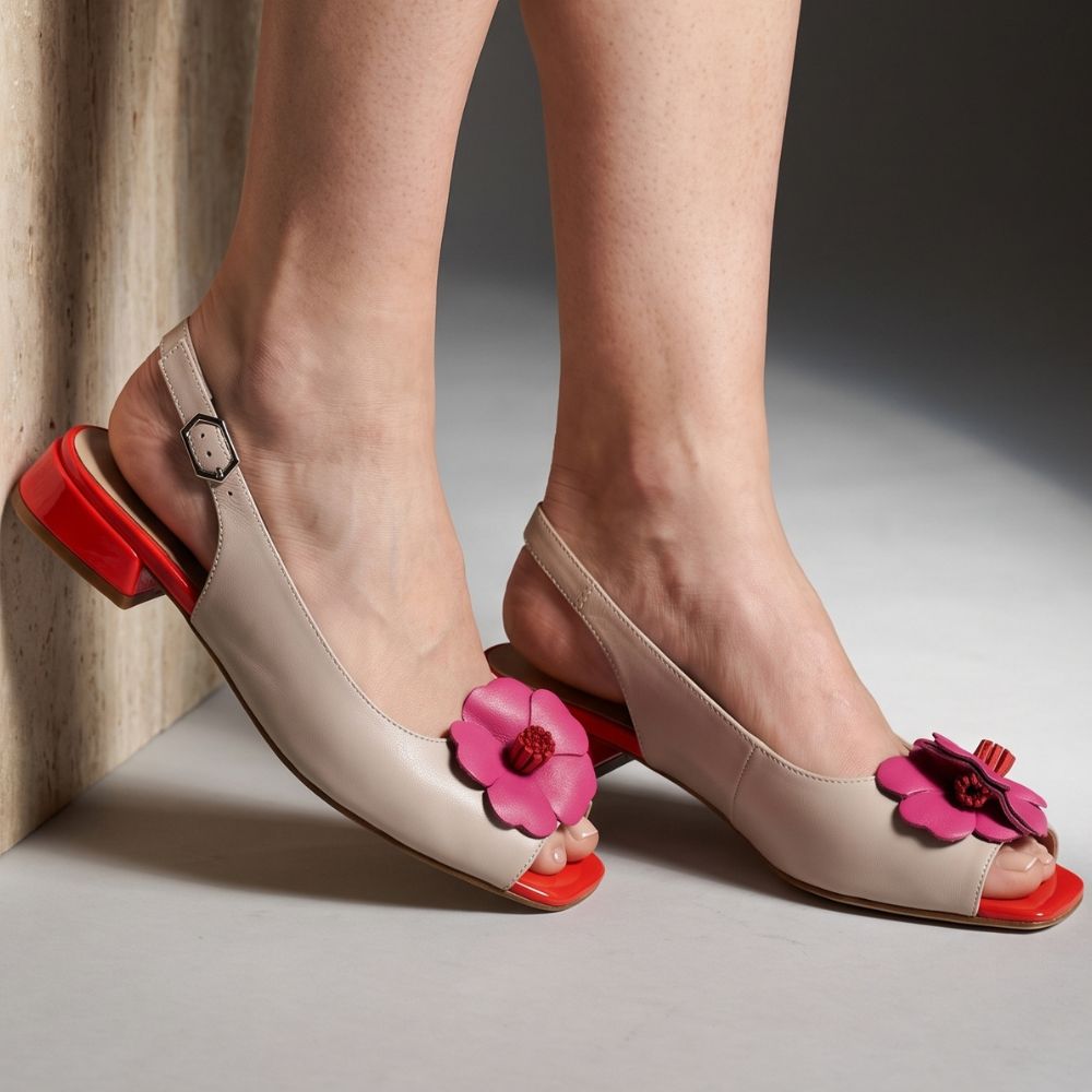 A close-up of a model's feet wearing beige peep-toe slingback sandals with bright pink flower embellishments and red square heels, set against a neutral studio background.