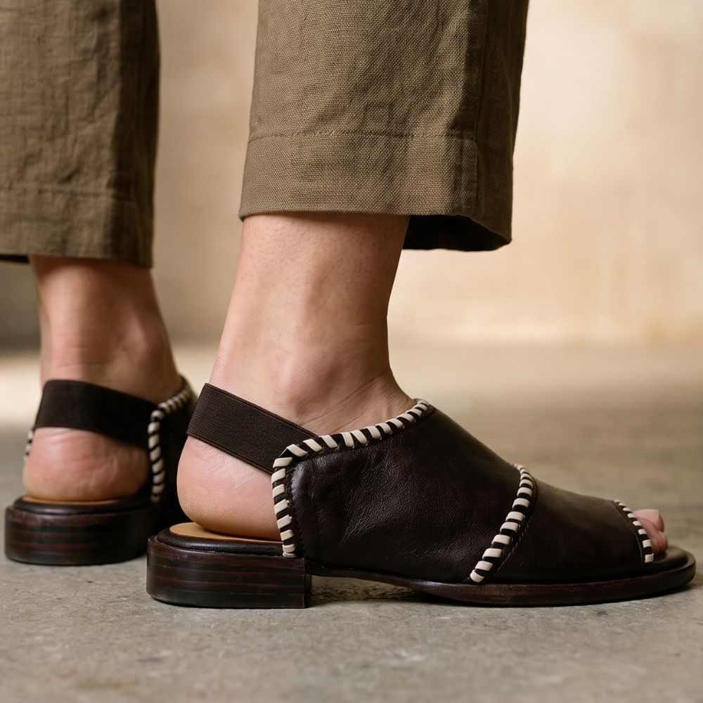 A close-up of a person wearing dark brown leather mule sandals with braided trim and stacked wooden-look soles, paired with brown trousers.