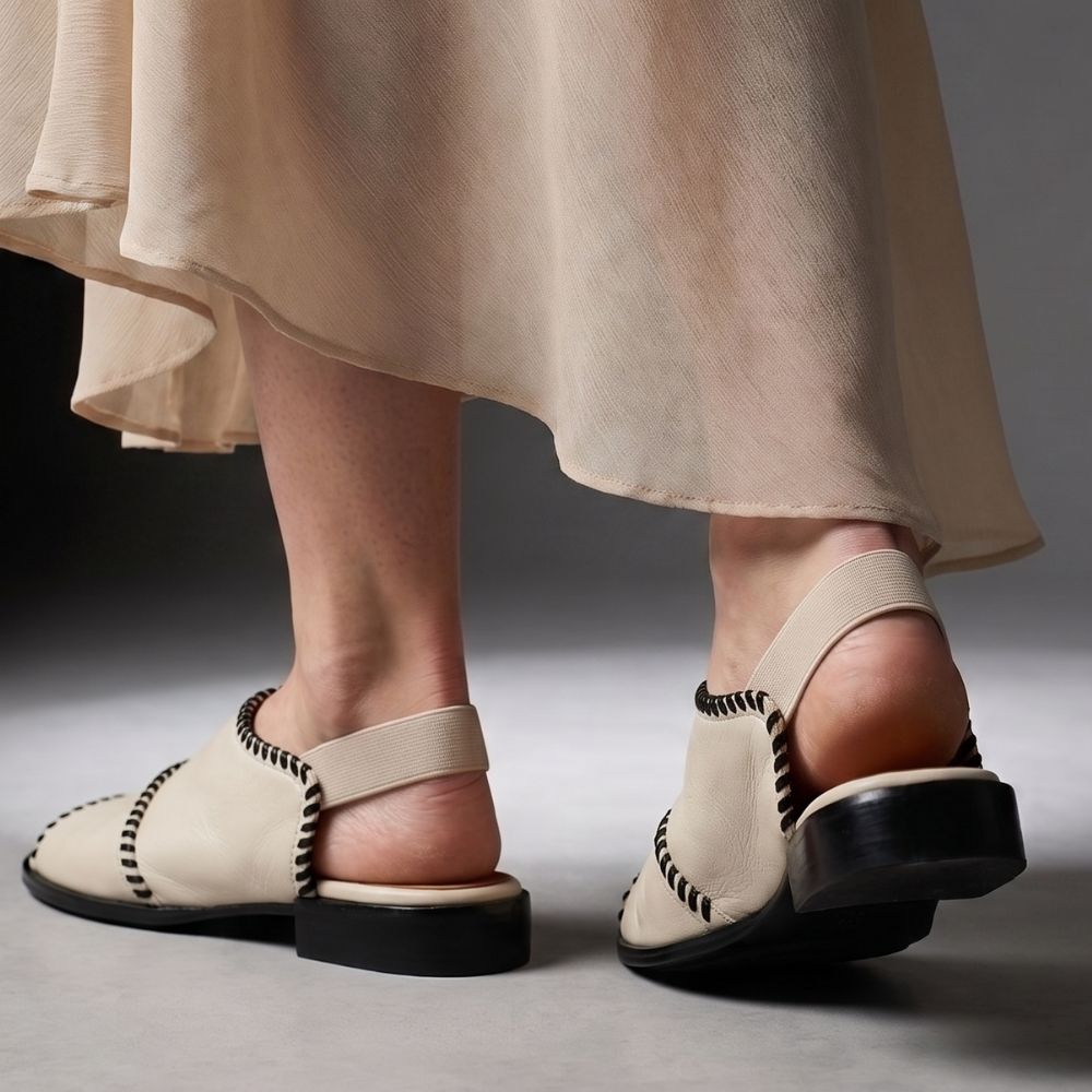 Cream leather slingback flat sandals with black braided trim and black soles, shown on a model's feet with a flowing beige skirt against a dark gray studio background.