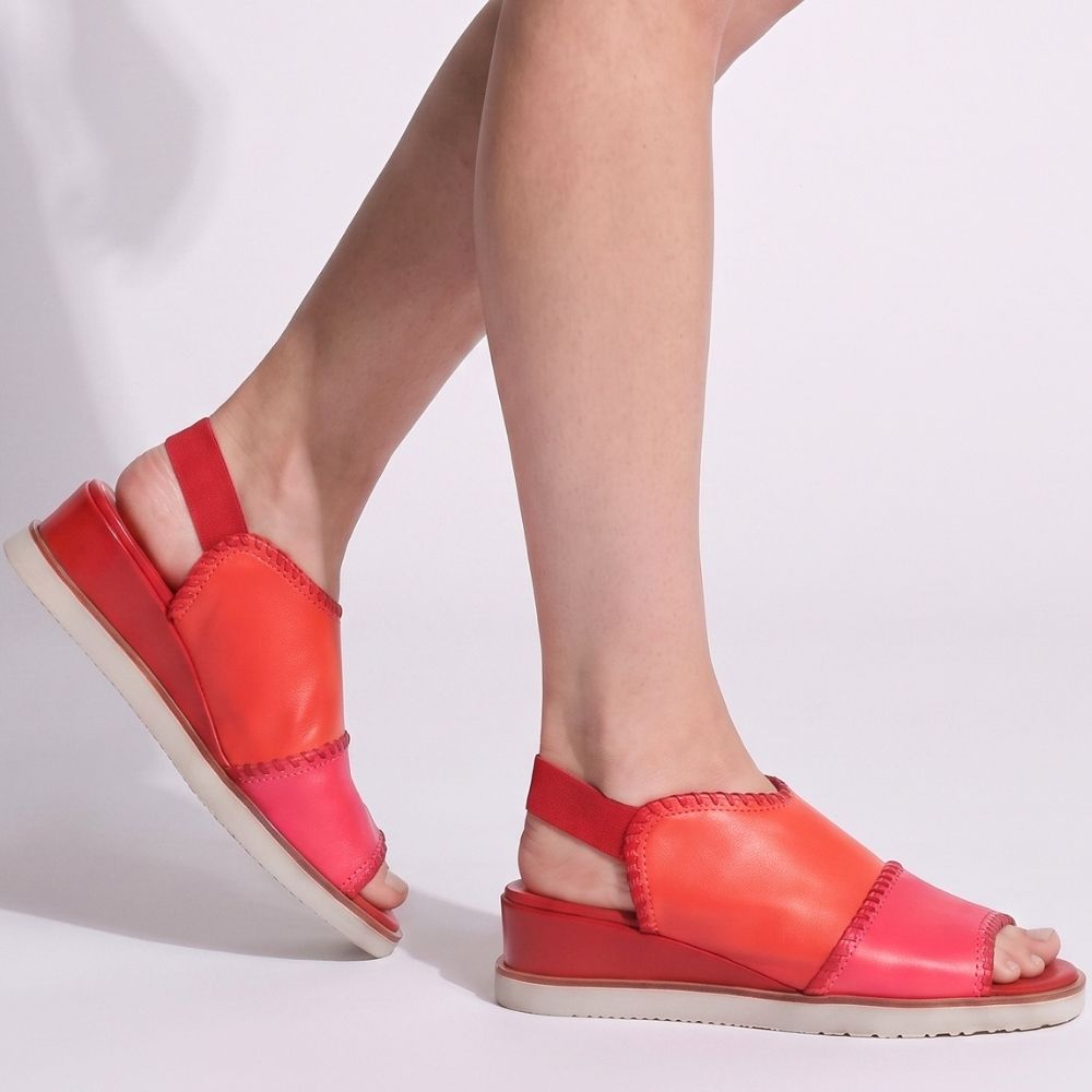 A pair of women's peep-toe wedge sandals in vibrant red, hot pink and orange tones, shown being worn on a model's feet against a clean white studio background.