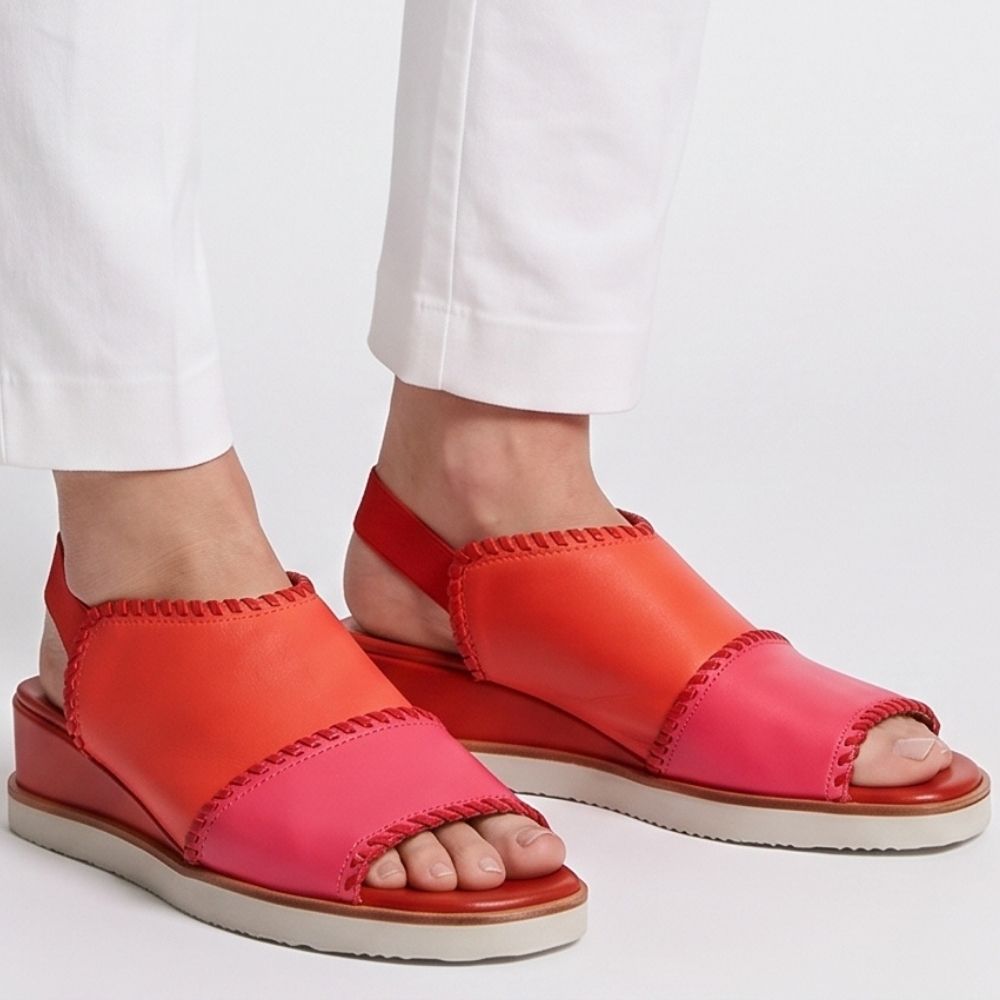 A pair of women's wedge sandals in vibrant shades of coral orange, hot pink, and red, shown on a model wearing white trousers against a clean white background.