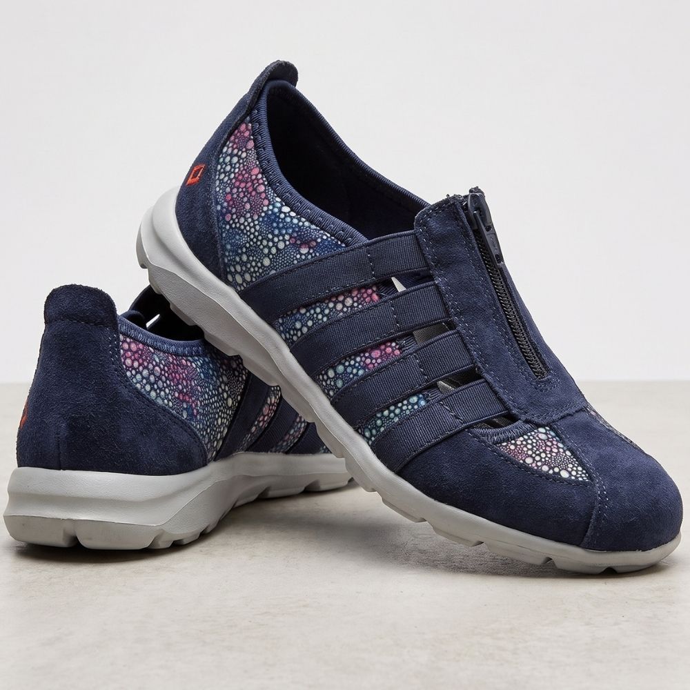 A pair of navy blue slip-on sneakers with a textured suede-like finish, featuring three elastic straps with colorful textile panels, a central zipper, and a thick white sole, shown against a neutral light gray background.