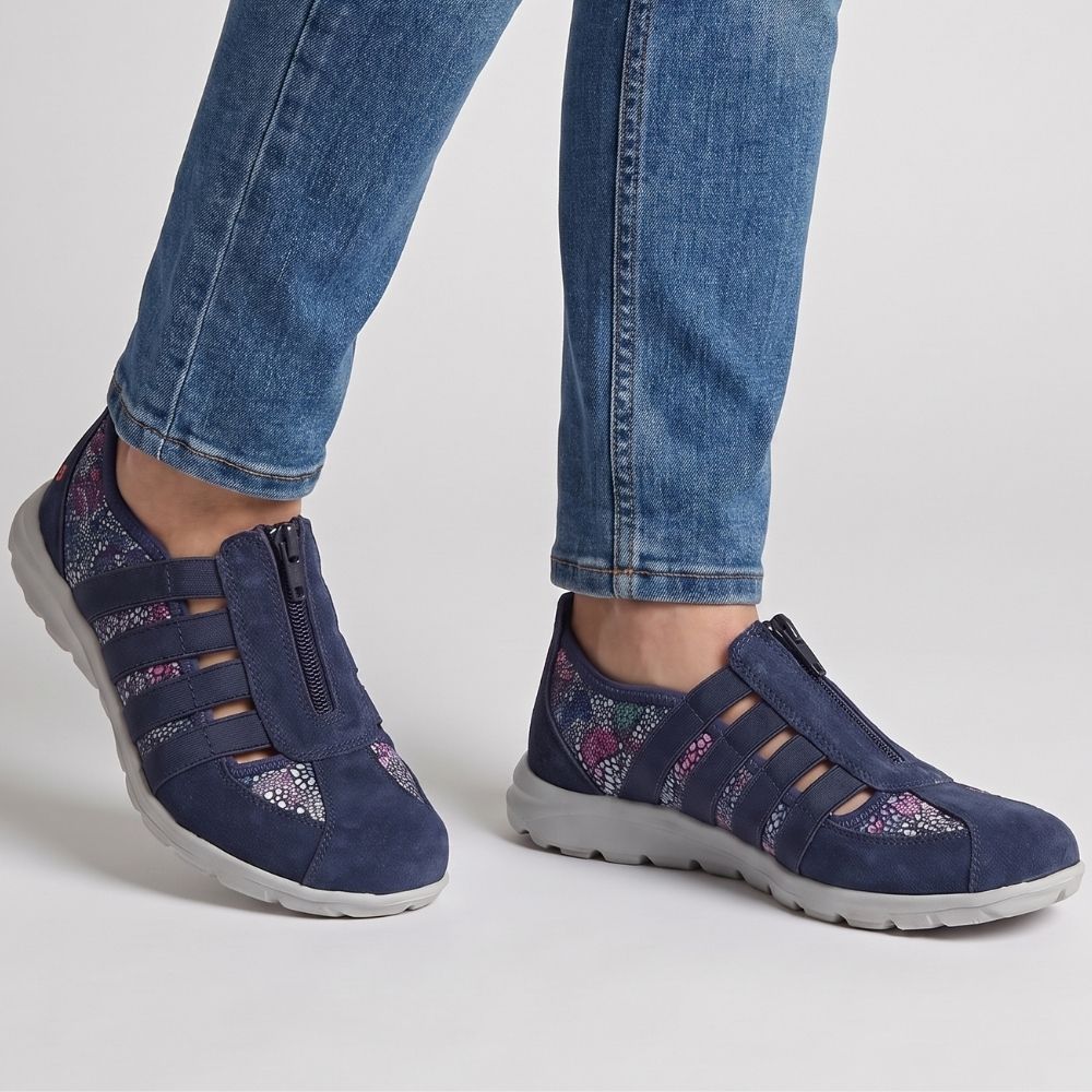 A person wearing blue jeans and navy slip-on suede sneakers with colourful patterned textile accents, photographed against a light gray studio background.