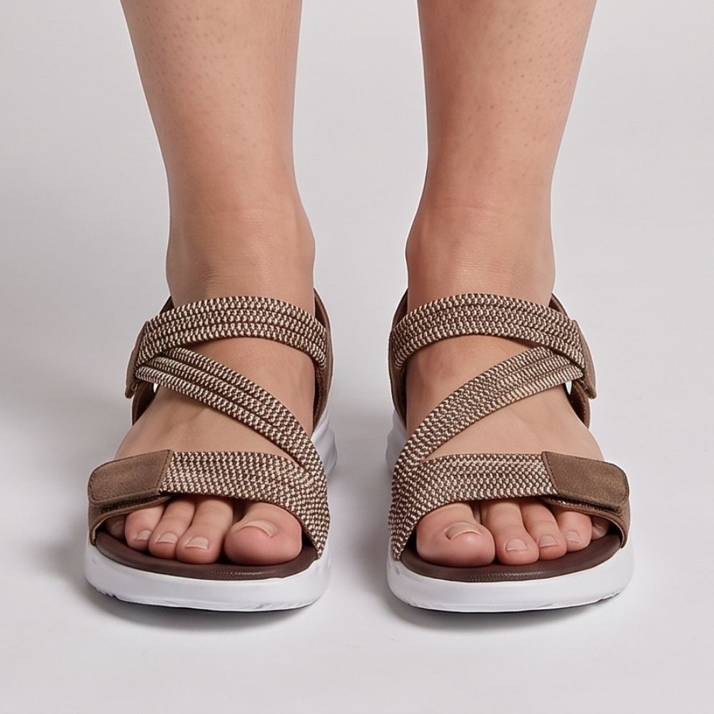 A person wearing dark brown leather and woven elastic strap sandals with white soles, shown in a front profile view against a white studio background.
