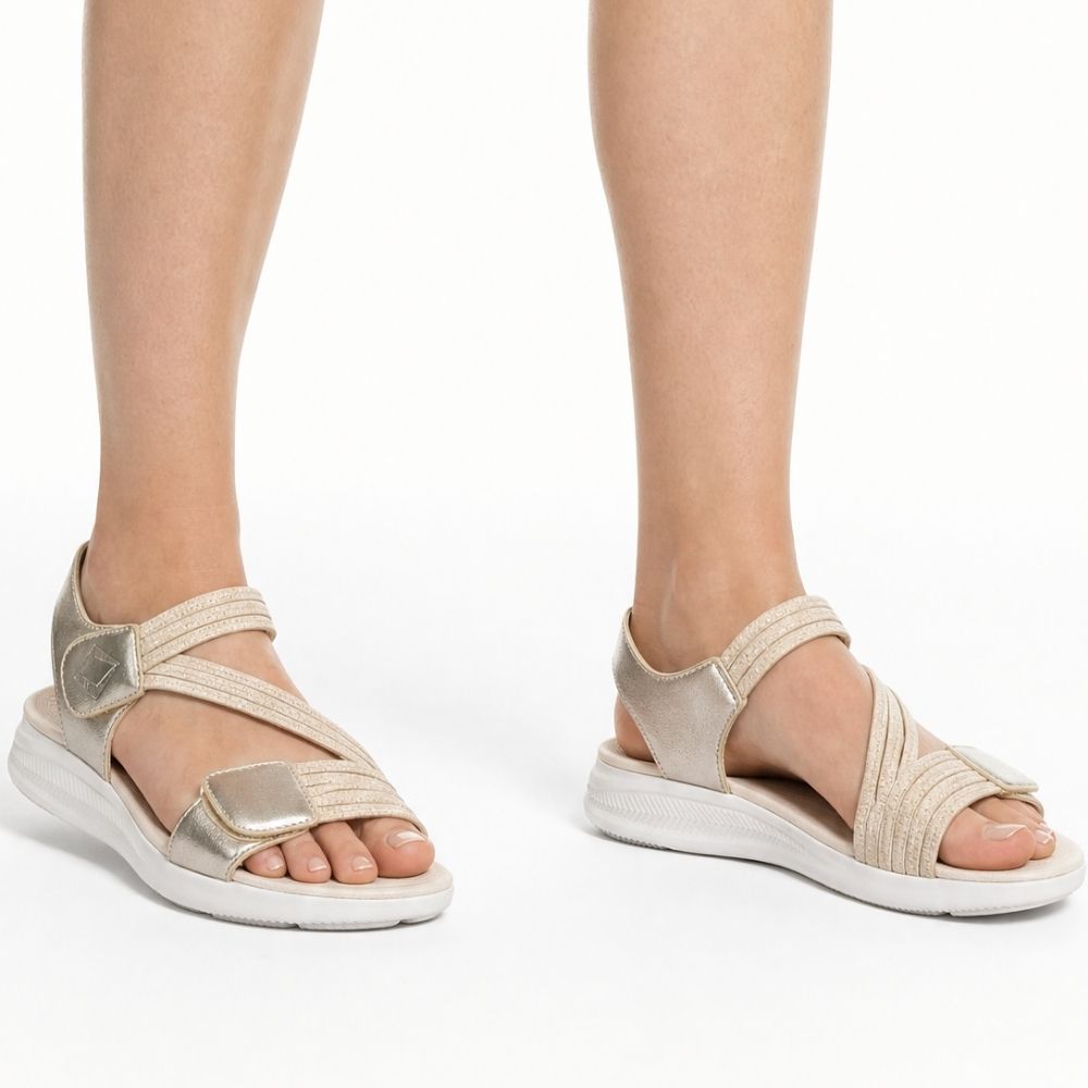 A person wearing a pair of metallic champagne gold sandals with multiple elastic woven straps and thick white cushioned soles, shown in a side profile on a studio background.