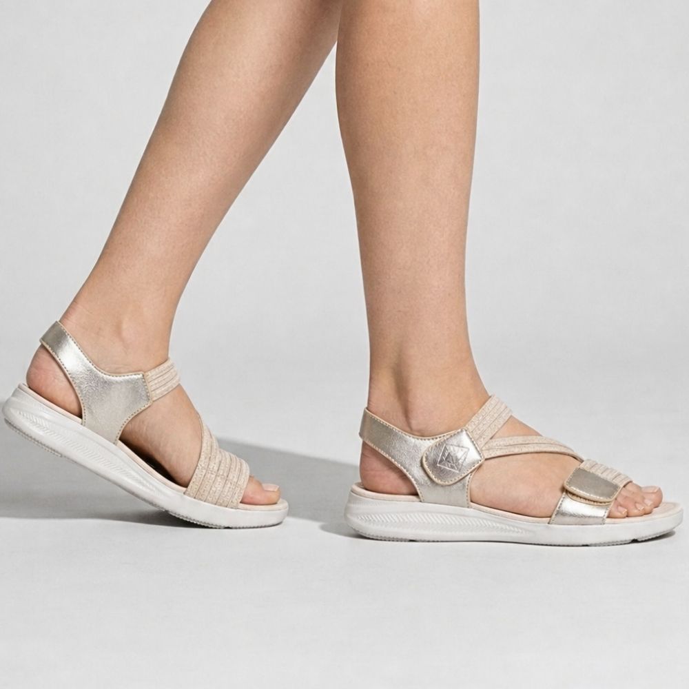A person wearing a pair of metallic champagne gold sandals with multiple elastic woven straps and thick white cushioned soles, shown in a side profile on a studio background.