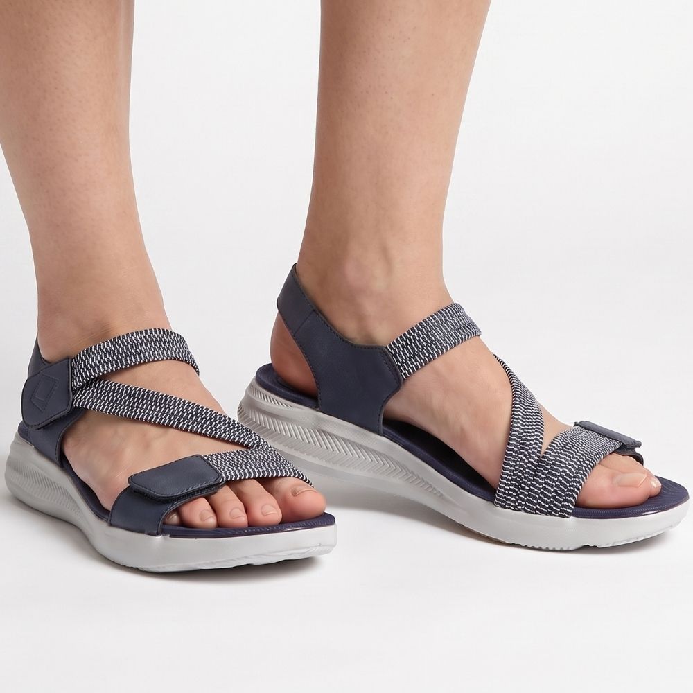 A person wearing navy blue sandals with elasticated navy and white straps on a white sole, standing against a studio background.