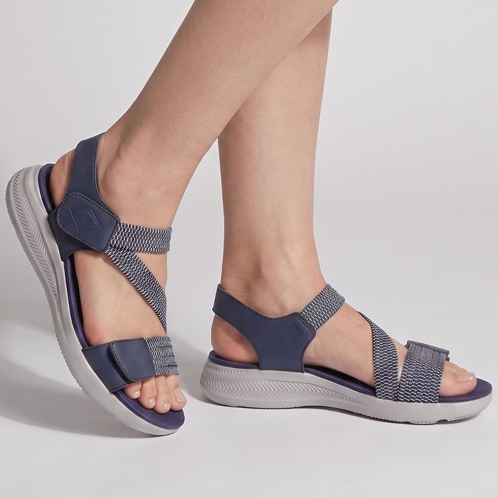 A person wearing navy blue sandals with elasticated navy and white straps on a white sole, standing against a studio background.