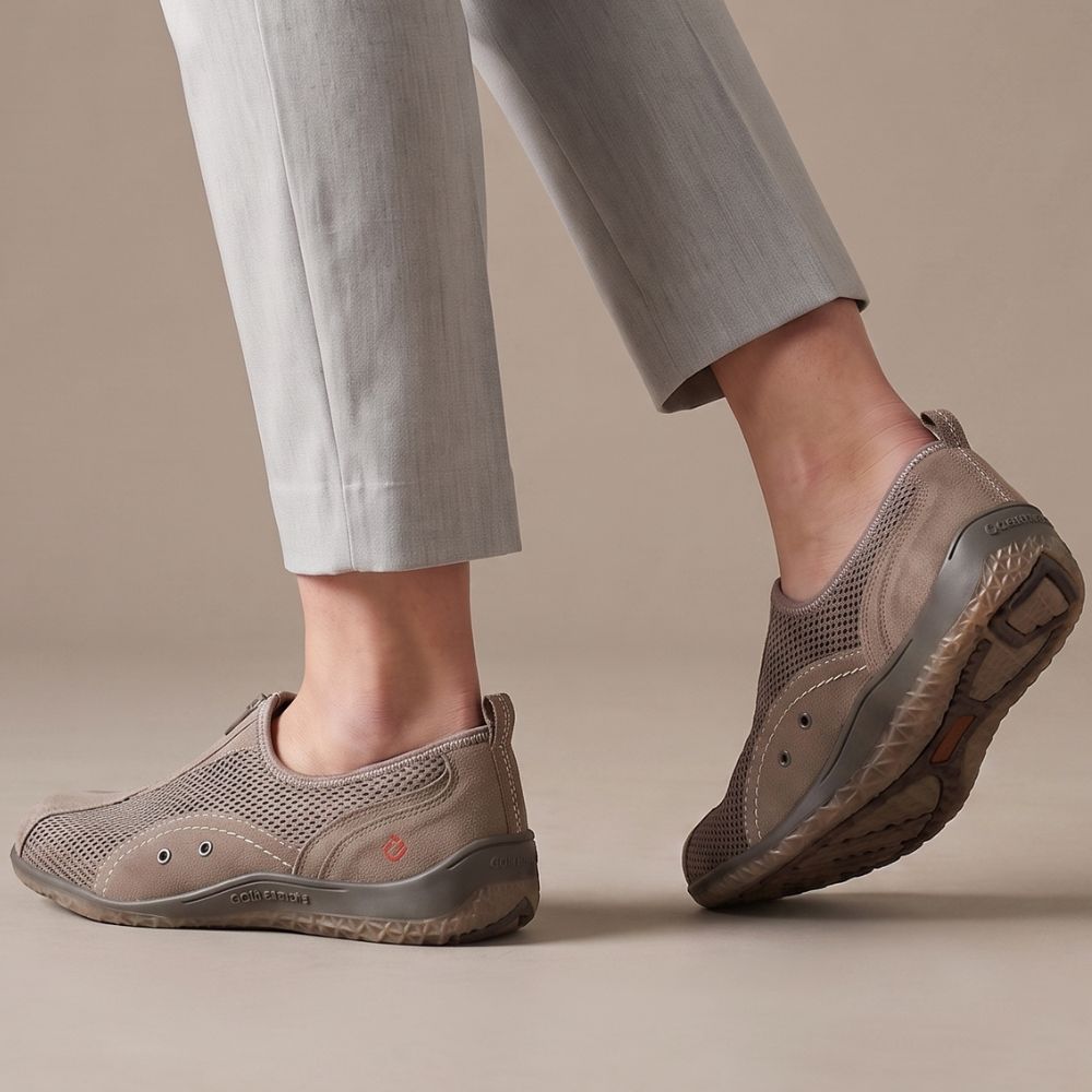 A person wearing taupe slip-on mesh sneakers with light gray trousers, shown from the calves down against a warm beige studio background.