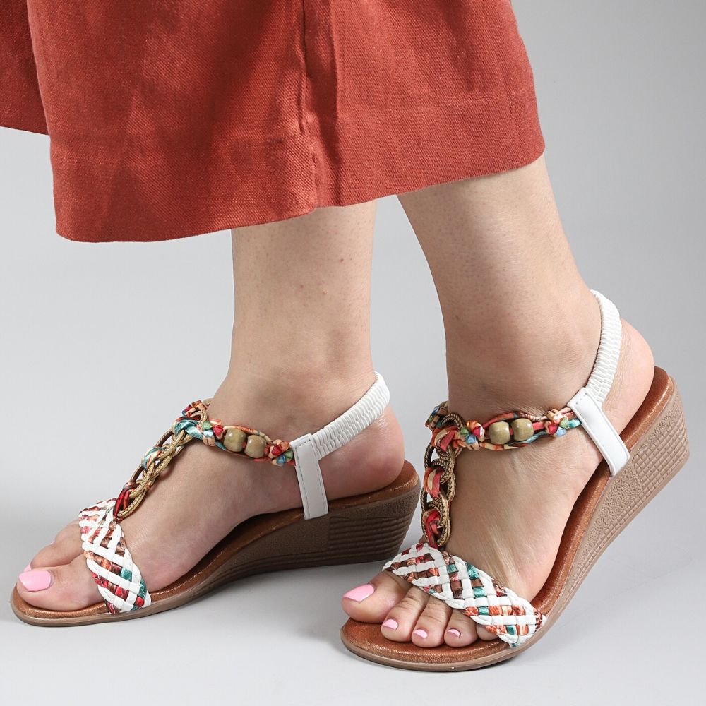 A women wearing a pair of wedge sandals with a multicolored woven and beaded T-strap upper, cushioned footbed, and a brown sole, against a grey studio background.