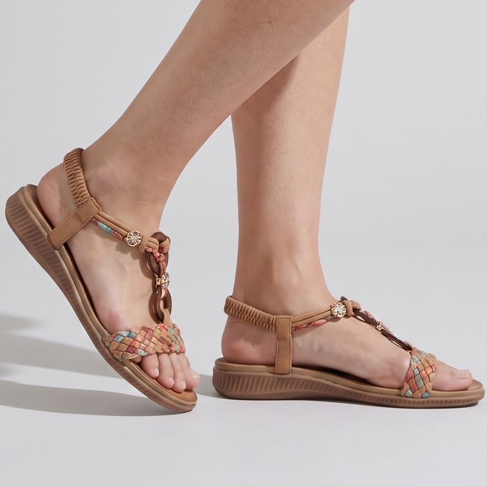 A pair of women's flat sandals shown on a model's feet against a light gray studio background. The sandals feature tan straps, a braided multicolor toe strap with small gold-tone charms, and a textured tan sole.
