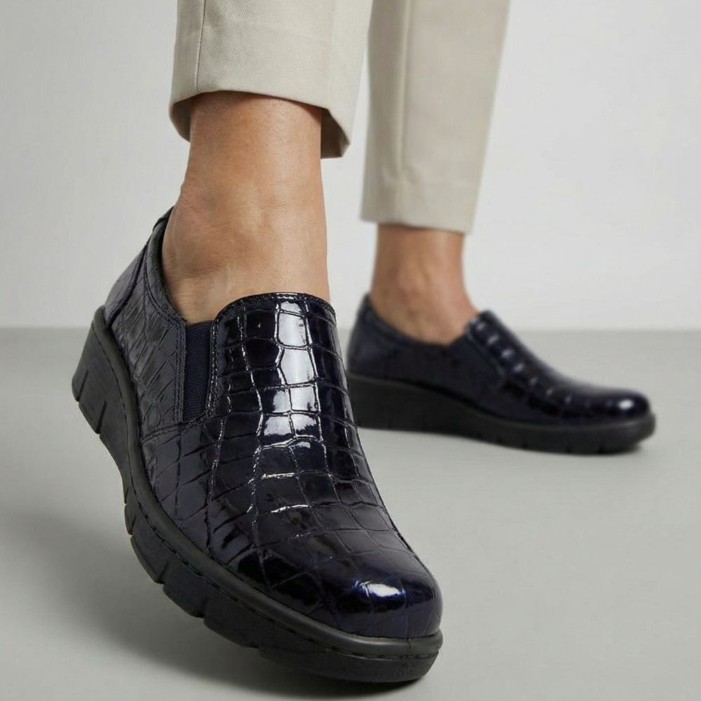 Black patent leather slip-on shoes with a textured pattern worn by a person on a light gray background
