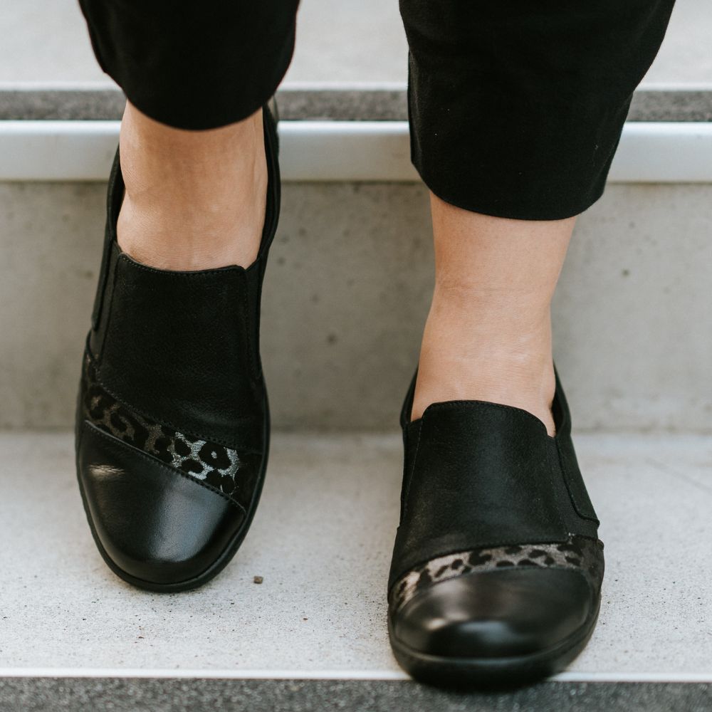 Black slip-on shoes with leopard print details worn by a person on concrete steps.