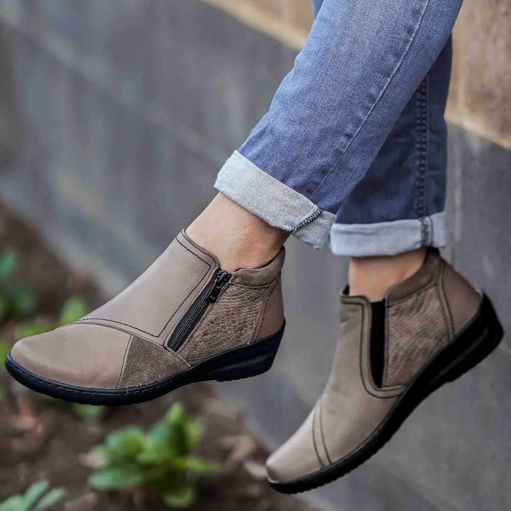 Person wearing taupe ankle boots with zippers, standing on a blurred natural background.