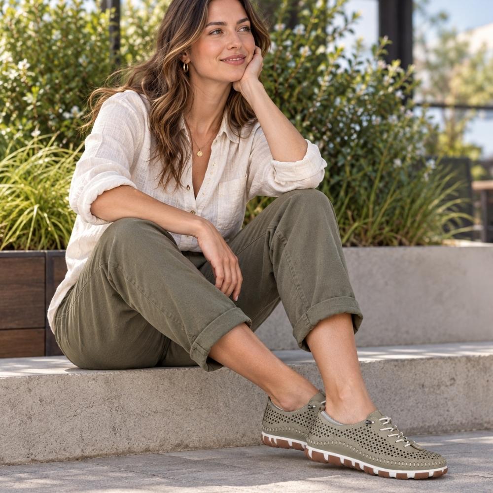 A lady wearing a linen top and pants sitting outside on a step, wearing a pair of khaki green perforated Turkish leather sneakers with bungee laces and brown and white soles.