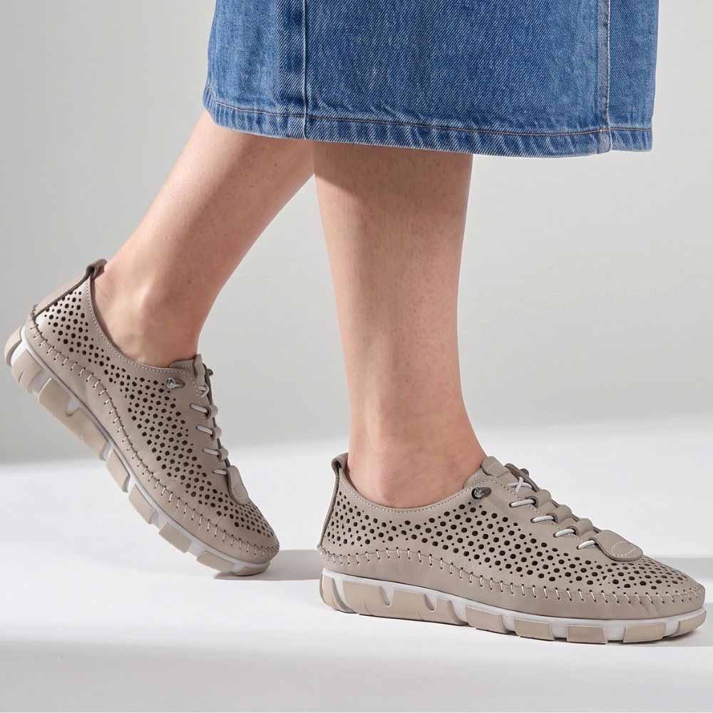 A lady wearing a pair of taupe perforated Turkish leather sneakers with bungee laces, stitched detailing and taupe and white soles, shown against a white background.