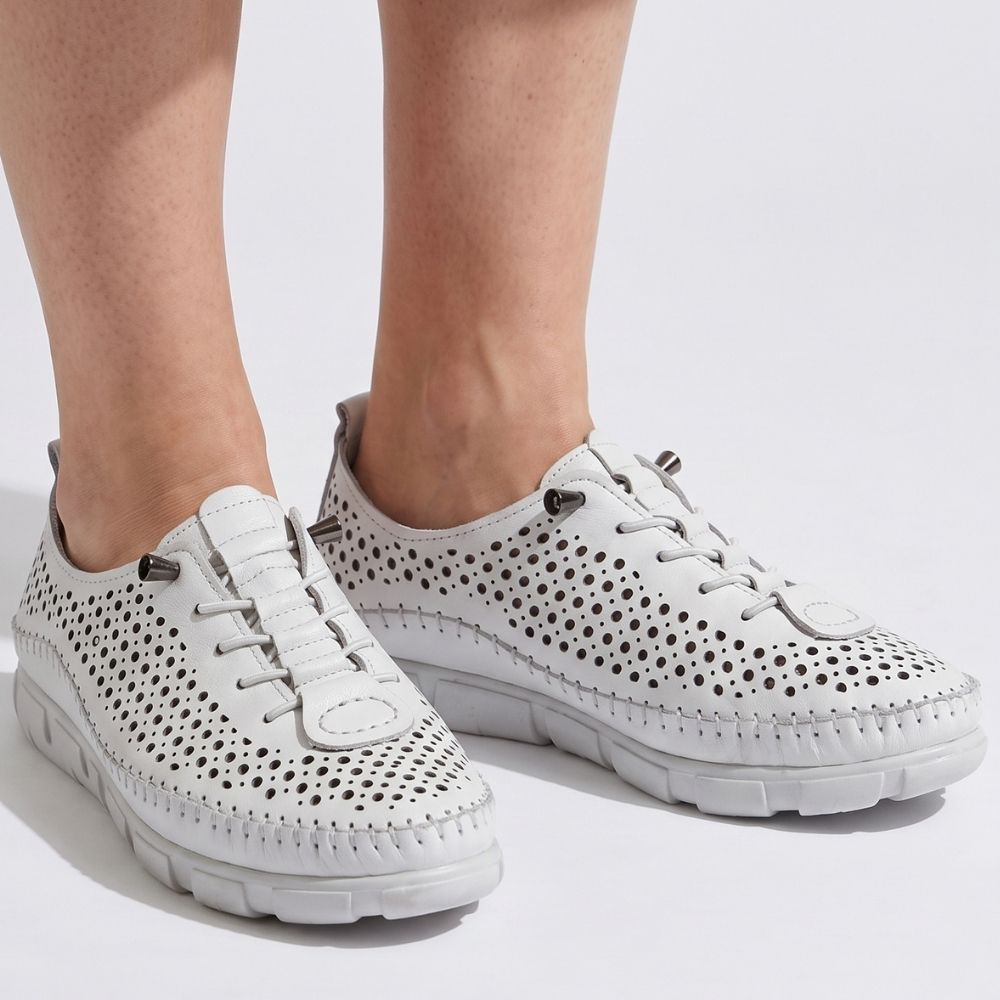 A lady in wearing a pair of white perforated Turkish leather sneakers with bungee laces, stitched detailing and a white sole, against a studio background.