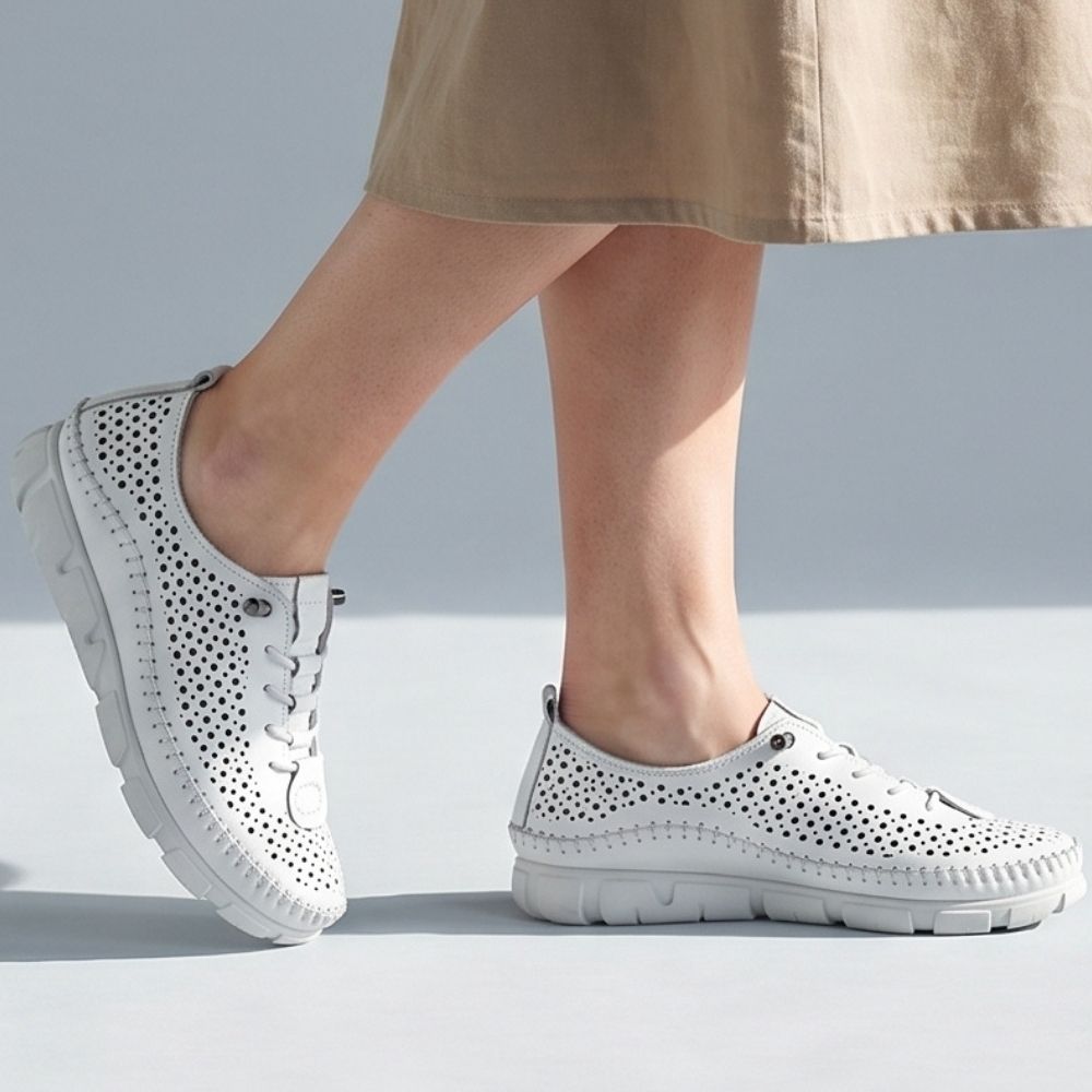 A lady in a linen skirt wearing a pair of white perforated Turkish leather sneakers with bungee laces, stitched detailing and a white sole, against a light blue studio background.