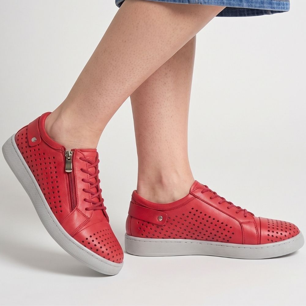 A pair of red perforated leather low-top sneakers with white soles, shown on a model wearing a blue denim skirt. The shoes feature side zippers, lace-up fronts, and metal details.