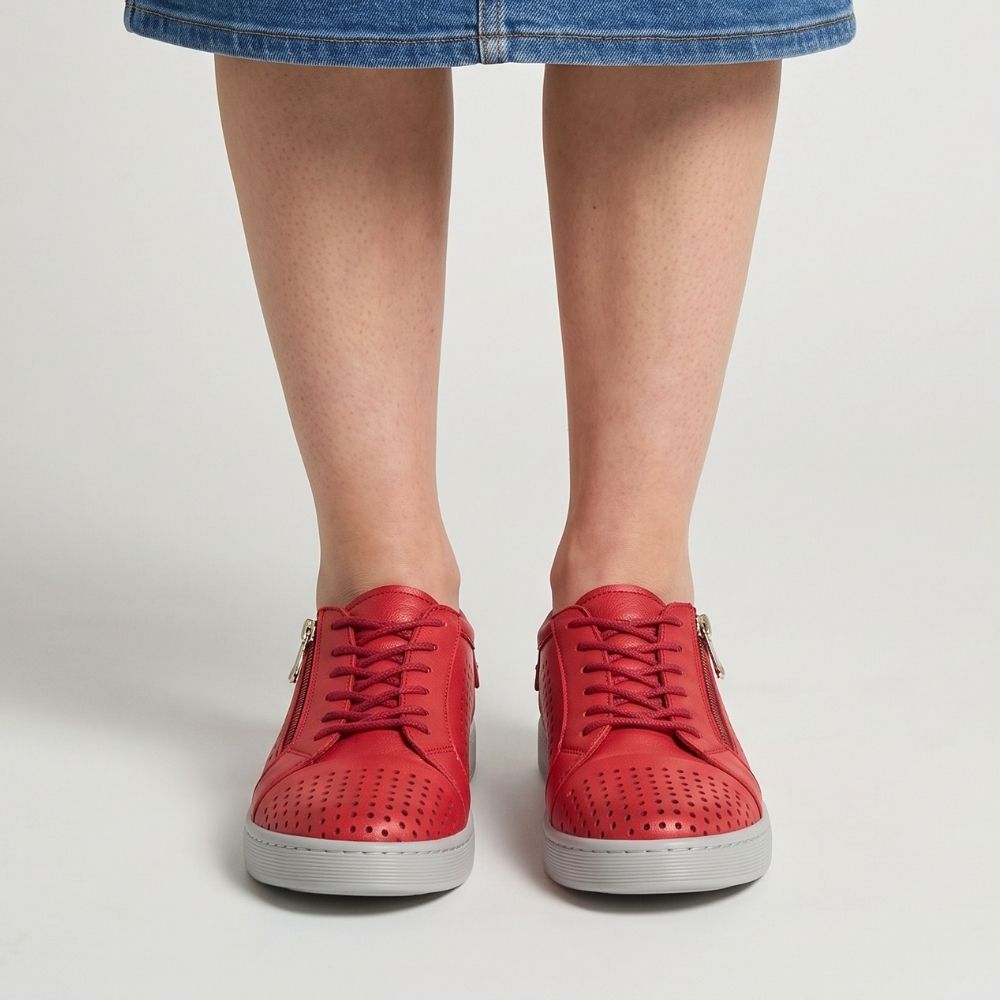 A pair of red perforated leather low-top sneakers with white soles, shown on a model wearing a blue denim skirt. The shoes feature side zippers, lace-up fronts, and metal details.