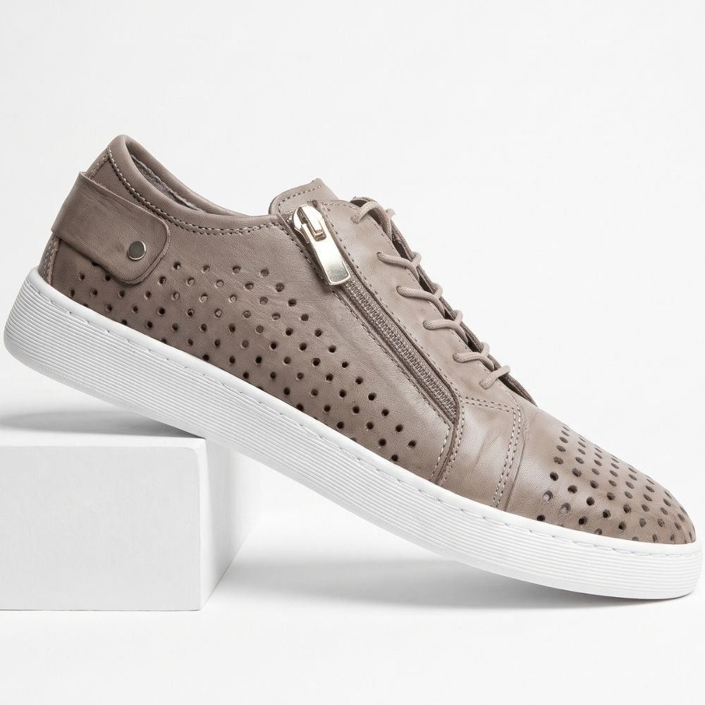 A taupe brown leather sneaker with perforated details, a silver-tone zipper, and a white rubber sole, shown resting on a white display block against a white background.