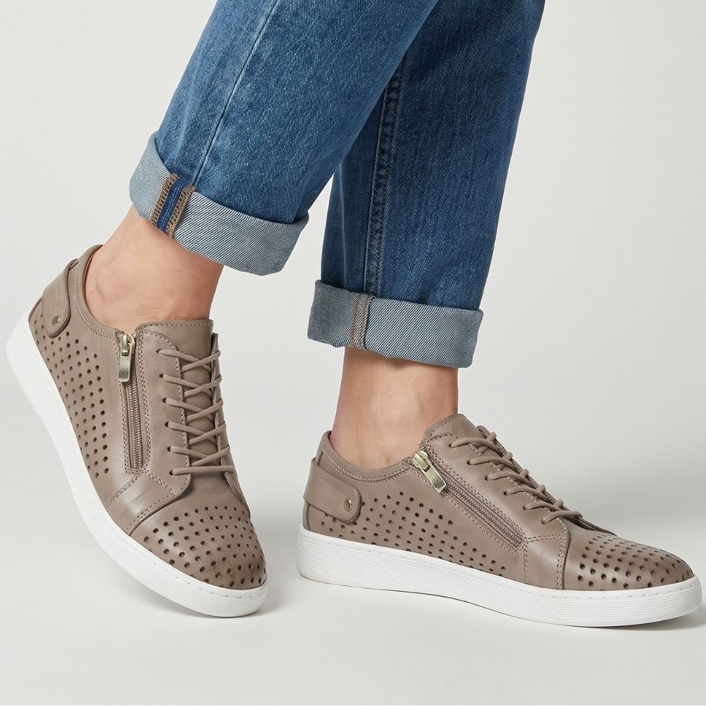 A pair of taupe perforated leather low-top sneakers with white soles, shown on a model wearing blue jeans. The shoes feature side zippers, lace-up fronts, and metal details.